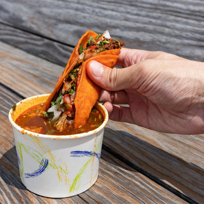 Birria taco being dipped into a rich red consommé made with Spicito Birria Bombs.