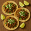 Three tacos with meat, onions, and cilantro on a wooden surface with lime wedges.